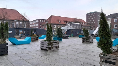 Auf dem Bild ist ein moderner, urbaner Platz mit gepflastertem Boden zu sehen. Im Vordergrund befinden sich blaue, geometrische Sitzmöbel und große Holzkübel mit grünen Nadelbäumen. Im Hintergrund stehen Gebäude mit roten Ziegeldächern. Ein schwarzer Container mit der Aufschrift „Hafen Büro“ und „Klima Kiste“ deutet auf eine kreative Nutzung des Raums hin. Die Stimmung wirkt ruhig und einladend bei leicht bewölktem Himmel.