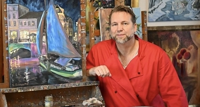 Ein Mann mit kurzem blond-grauem Haar und Bart sitzt in entspannter Haltung in einem Atelier vor mehreren farbenfrohen Gemälden. Er trägt einen leuchtend roten Arbeitskittel über einem hellen Shirt und blickt freundlich in die Kamera. Neben ihm steht auf einer Staffelei ein Bild mit einer nächtlichen Hafen- oder Altstadtszene in Blau-, Lila- und Rosatönen, auf dem ein Segelboot zu sehen ist. Im Hintergrund hängen weitere Kunstwerke in warmen und kühlen Farben an Backsteinwänden und Holzstaffeleien. Die Stimmung wirkt kreativ, ruhig und einladend.