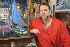 Ein Mann mit kurzem blond-grauem Haar und Bart sitzt in entspannter Haltung in einem Atelier vor mehreren farbenfrohen Gemälden. Er trägt einen leuchtend roten Arbeitskittel über einem hellen Shirt und blickt freundlich in die Kamera. Neben ihm steht auf einer Staffelei ein Bild mit einer nächtlichen Hafen- oder Altstadtszene in Blau-, Lila- und Rosatönen, auf dem ein Segelboot zu sehen ist. Im Hintergrund hängen weitere Kunstwerke in warmen und kühlen Farben an Backsteinwänden und Holzstaffeleien. Die Stimmung wirkt kreativ, ruhig und einladend.