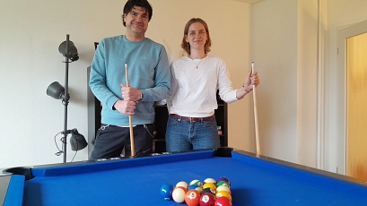 Zwei Personen stehen hinter einem blauen Billardtisch in einem hellen Aufenthaltsraum und halten jeweils ein Billardqueue in der Hand. Auf dem Tisch liegen farbige Billardkugeln in Dreiecksformation. Der Mann links trägt einen hellblauen Pullover und dunkle Hose, die Frau rechts ein weißes Langarmshirt und blaue Jeans. Beide lächeln in die Kamera. Im Hintergrund stehen eine schwarze Stehlampe und ein Regal vor einer weißen Wand. Die Stimmung wirkt entspannt, freundlich und einladend.