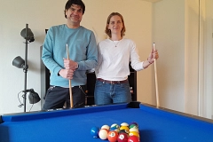 Zwei Personen stehen hinter einem blauen Billardtisch in einem hellen Aufenthaltsraum und halten jeweils ein Billardqueue in der Hand. Auf dem Tisch liegen farbige Billardkugeln in Dreiecksformation. Der Mann links trägt einen hellblauen Pullover und dunkle Hose, die Frau rechts ein weißes Langarmshirt und blaue Jeans. Beide lächeln in die Kamera. Im Hintergrund stehen eine schwarze Stehlampe und ein Regal vor einer weißen Wand. Die Stimmung wirkt entspannt, freundlich und einladend.