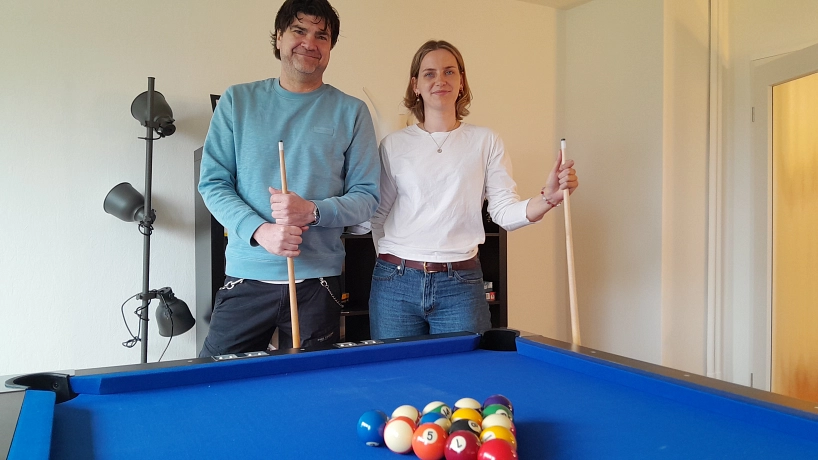 Eröffnung Jugendhaus Bützfleth: Luisa Maibohm und Marc Olszewski Zwei Personen stehen hinter einem blauen Billardtisch in einem hellen Aufenthaltsraum und halten jeweils ein Billardqueue in der Hand. Auf dem Tisch liegen farbige Billardkugeln in Dreiecksformation. Der Mann links trägt einen hellblauen Pullover und dunkle Hose, die Frau rechts ein weißes Langarmshirt und blaue Jeans. Beide lächeln in die Kamera. Im Hintergrund stehen eine schwarze Stehlampe und ein Regal vor einer weißen Wand. Die Stimmung wirkt entspannt, freundlich und einladend. © Hansestadt Stade
