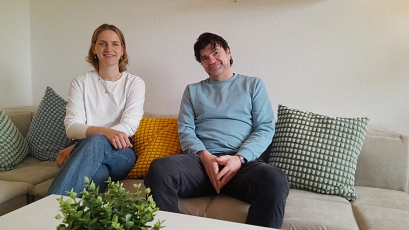 Zwei Personen sitzen nebeneinander auf einem hellen Sofa in einem gemütlich eingerichteten Aufenthaltsraum. Die Frau trägt ein weißes Langarmshirt und blaue Jeans, der Mann einen hellblauen Pullover und dunkle Hose. Mehrere strukturierte Kissen in Grün- und Gelbtönen sorgen für farbliche Akzente. Im Vordergrund steht ein kleiner Tisch mit einer grünen Topfpflanze. Der Raum ist hell und modern gestaltet, die Atmosphäre ruhig, offen und wohnlich.