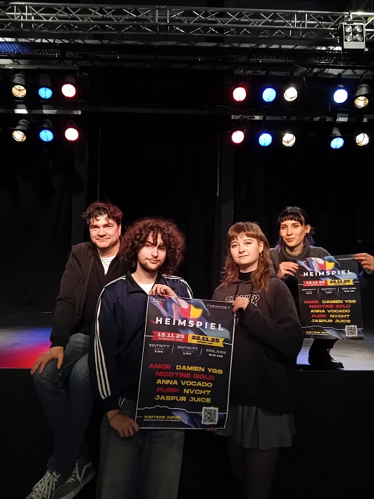 Vier junge Erwachsene stehen auf einer dunklen Bühne mit schwarzem Hintergrund und halten farbige Veranstaltungsplakate in den Händen. Die Plakate bewerben das Musikfestival „Heimspiel“ im Alten Schlachthof Stade. Die Szene ist von Scheinwerfern in Blau, Rot und Weiß beleuchtet, was eine lebendige und zugleich konzentrierte Atmosphäre schafft. Links sitzt eine Person im schwarzen Hoodie und Jeans auf der Bühnenkante, daneben stehen zwei Personen: eine mit lockigem Haar und Sportjacke, die andere mit Ponyfrisur, Sweatshirt und Faltenrock. Ganz rechts steht eine weitere Person mit dunklem Pony und dunklem Outfit, halb im Bühnenhintergrund. Die Stimmung ist konzentriert und engagiert.