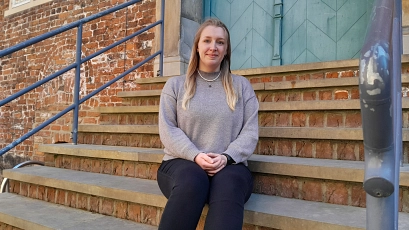 Eine junge Frau mit langen, blondierten Haaren sitzt auf einer Backsteintreppe vor einer historischen Gebäudefassade mit grünlicher Tür. Sie trägt einen grauen Pullover und blickt freundlich in die Kamera. Metallgeländer rahmen die Treppe ein.