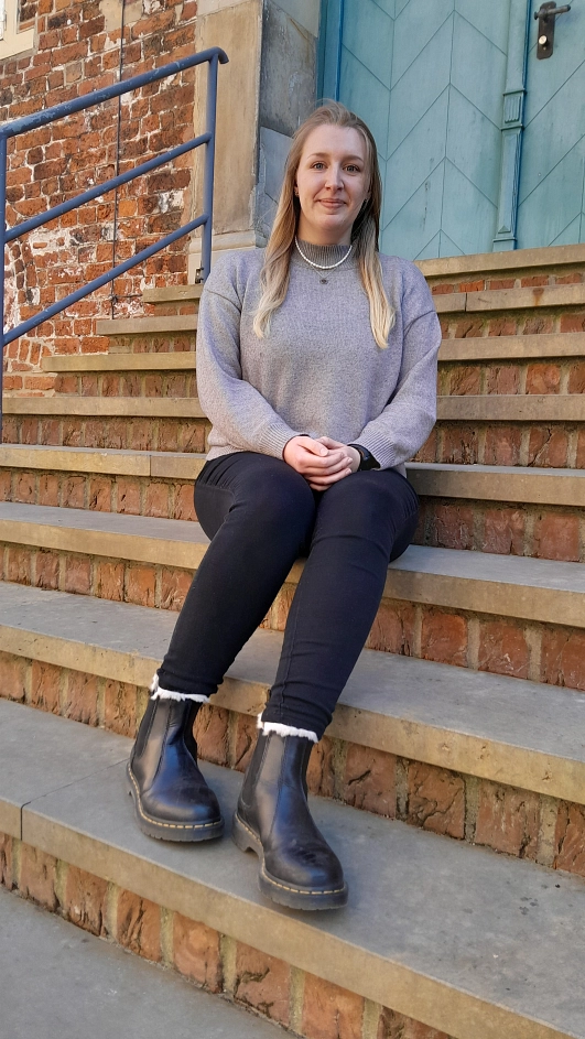 Eine junge Frau mit langen, blondierten Haaren sitzt auf einer Backsteintreppe vor einer historischen Gebäudefassade mit grünlicher Tür. Sie trägt einen grauen Pullover und blickt freundlich in die Kamera. Metallgeländer rahmen die Treppe ein.