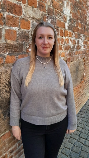 Eine junge Frau mit langen, glatten, blondierten Haaren steht vor einer alten Backsteinmauer in der Sonne. Sie trägt einen grauen Pullover, eine schwarze Hose und schaut freundlich in die Kamera. Der Boden ist mit Kopfsteinpflaster bedeckt.