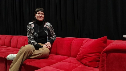 Eine Frau, Catherine Bartl, sitzt auf einem langen, roten Sofa vor einem schwarzen Vorhang. Sie trägt eine gemusterte Jacke, ein schwarzes Oberteil und beigefarbene Hosen. Die entspannte Atmosphäre vermittelt einen einladenden Eindruck, der Raum wirkt modern und gemütlich.