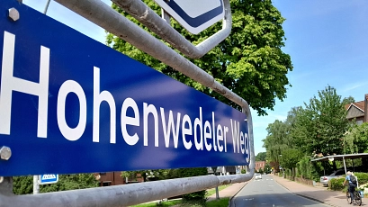 Straßennamensschild des Hohenwedeler Weges in Großaufnahme. Das Schild führt den Blick von links ins Bild hinein. Im Hintergrund ist die Straße zu sehen, auf deren Fußweg ein Radfahrer unterwegs ist