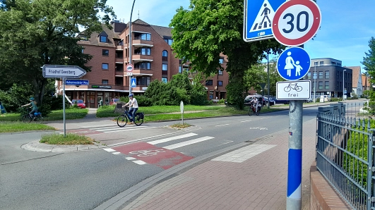 Einmündungsbereich des Hohenwedeler Weges. Im Vordergrund ist ein Schild zu sehen, dass auf Tempo 30 und die Möglichkeit für Radfahrende hinweist, den Gehweg zu benutzen. Im Hintergrund sind Radfahrende auf der Straße zu sehen