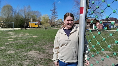 Eine junge Frau steht lächelnd an einem Fußballtor auf einem weitläufigen Spiel- und Bolzplatz. Im Hintergrund sind Spielgeräte, eine gelbe Bauwagenhütte, Container und Gebäude zu sehen. Der Himmel ist blau, es ist sonnig.