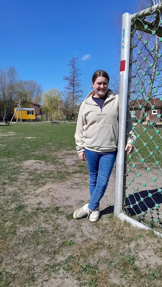 Eine junge Frau steht lächelnd an einem Fußballtor auf einem weitläufigen Spiel- und Bolzplatz. Im Hintergrund sind Spielgeräte, eine gelbe Bauwagenhütte, Container und Gebäude zu sehen. Der Himmel ist blau, es ist sonnig.