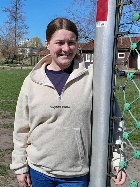Eine junge Frau mit zusammengebundenen braunen Haaren steht lächelnd an einem Fußballtor auf einem Bolzplatz. Sie trägt eine helle Fleecejacke mit der Aufschrift „Originals Studio“ und blaue Jeans. Im Hintergrund sind grüne Wiese, Bäume und Gebäude zu sehen.