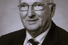 Hans-Hermann Ott Schwarz-weißes Porträt eines älteren Mannes mit kurzem, lichtem Haar und Brille, der leicht seitlich zur Kamera blickt. Er trägt einen dunklen Anzug, ein helles Hemd sowie eine gestreifte Krawatte. Am Revers ist ein kleines Abzeichen angebracht. Der Hintergrund ist neutral und weich verlaufend in Grautönen gehalten. Die Stimmung wirkt ruhig, seriös und würdevoll, passend zu einem offiziellen Porträt.