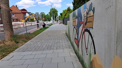 Ein frisch gepflasterter Gehweg in einer Wohnstraße mit angrenzender Baustelle. Links fährt eine Person auf einem Fahrrad den roten Radweg entlang. Rechts ziert ein farbenfrohes Wandbild aus Graffiti die Mauer – es zeigt ein rotes Fahrrad mit Einkaufskorb, darin Gemüse. Im Hintergrund sind Baustellenfahrzeuge, Verkehrsschilder und Absperrgitter zu sehen.