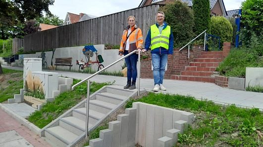 Zwei Personen in Sicherheitskleidung stehen auf einem gepflasterten Weg vor einer neu gestalteten Treppenanlage mit Edelstahlgeländer. Im Hintergrund ist eine mit farbenfrohen Graffiti bemalte Betonwand zu sehen – dargestellt ist eine Person auf einem roten Fahrrad. Links im Bild sind ebenfalls bemalte Versorgungskästen und weitere Motive sichtbar.