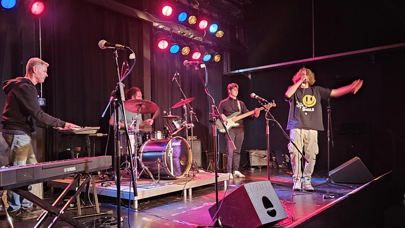 Heimspiel-Workshop 2025 Eine vierköpfige Band steht auf einer Bühne in farbigem Scheinwerferlicht. Links spielt ein Musiker Keyboard, daneben sitzt ein Schlagzeuger am Drumset. In der Mitte spielt ein Bassist, rechts singt ein Sänger mit lockigem Haar und ausdrucksstarker Gestik ins Mikrofon. Im Vordergrund steht weiteres Bühnenequipment, unter anderem ein Yamaha-Keyboard. © Hansestadt Stade