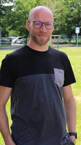 Ein Mann mit Glatze, Lukas Funke, und kurzem Bart steht lächelnd im Freien. Er trägt eine runde Brille und ein T-Shirt mit schwarzem oberen Teil und grauem unteren Bereich sowie einer grauen Brusttasche. Im Hintergrund sind eine grüne Wiese, Bäume und unscharf parkende Autos zu sehen.
