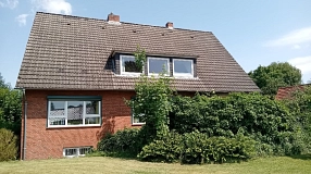 Ein großes Wohnhaus aus roten Backsteinen steht an einer gepflegten Grünfläche unter blauem Himmel mit wenigen Wolken. Das Haus besitzt ein steiles, dunkel gedecktes Satteldach mit mehreren Dachfenstern und weißen Fensterrahmen. Vor der rechten Haushälfte wachsen dichte grüne Sträucher und Kletterpflanzen, die einen Teil der Fassade bedecken. Im Hintergrund sind weitere Bäume und Dächer benachbarter Häuser zu sehen. Die sommerliche Aufnahme wirkt ruhig, freundlich und wohnlich.&nbsp;&copy;&nbsp;Hansestadt Stade