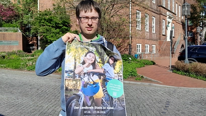 Eine Person steht im Freien auf einem gepflasterten Platz und hält ein großes, leicht geknicktes Plakat zum Stadtradeln in die Kamera. Sie trägt eine blaue Jeans, dunkle Schuhe sowie einen blau-grünen Hoodie unter einer Jacke und hat eine Brille. Neben ihr steht ein Fahrrad, dessen Vorderrad und Lenker im Vordergrund sichtbar sind. Das Plakat zeigt zwei lachende Kinder mit Fahrrädern in warmem Sonnenlicht. Dominierende Farben sind Grün- und Gelbtöne im Hintergrund sowie Blau beim Fahrradhelm. Ein grüner Kreis hebt einen Hinweis zur App hervor. Im Hintergrund des Fotos sind ein rotes Backsteingebäude, ein parkendes dunkles Auto, Bäume mit frischem Grün und ein blauer Himmel zu sehen. Das Licht ist hell und sonnig, die Stimmung insgesamt freundlich, aktiv und einladend.