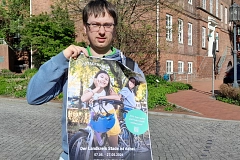 Eine Person steht im Freien auf einem gepflasterten Platz und hält ein großes, leicht geknicktes Plakat zum Stadtradeln in die Kamera. Sie trägt eine blaue Jeans, dunkle Schuhe sowie einen blau-grünen Hoodie unter einer Jacke und hat eine Brille. Neben ihr steht ein Fahrrad, dessen Vorderrad und Lenker im Vordergrund sichtbar sind. Das Plakat zeigt zwei lachende Kinder mit Fahrrädern in warmem Sonnenlicht. Dominierende Farben sind Grün- und Gelbtöne im Hintergrund sowie Blau beim Fahrradhelm. Ein grüner Kreis hebt einen Hinweis zur App hervor. Im Hintergrund des Fotos sind ein rotes Backsteingebäude, ein parkendes dunkles Auto, Bäume mit frischem Grün und ein blauer Himmel zu sehen. Das Licht ist hell und sonnig, die Stimmung insgesamt freundlich, aktiv und einladend.