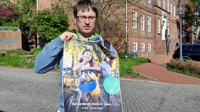 Eine Person steht im Freien auf einem gepflasterten Platz und hält ein großes, leicht geknicktes Plakat zum Stadtradeln in die Kamera. Sie trägt eine blaue Jeans, dunkle Schuhe sowie einen blau-grünen Hoodie unter einer Jacke und hat eine Brille. Neben ihr steht ein Fahrrad, dessen Vorderrad und Lenker im Vordergrund sichtbar sind. Das Plakat zeigt zwei lachende Kinder mit Fahrrädern in warmem Sonnenlicht. Dominierende Farben sind Grün- und Gelbtöne im Hintergrund sowie Blau beim Fahrradhelm. Ein grüner Kreis hebt einen Hinweis zur App hervor. Im Hintergrund des Fotos sind ein rotes Backsteingebäude, ein parkendes dunkles Auto, Bäume mit frischem Grün und ein blauer Himmel zu sehen. Das Licht ist hell und sonnig, die Stimmung insgesamt freundlich, aktiv und einladend.&nbsp;&copy;&nbsp;Hansestadt Stade