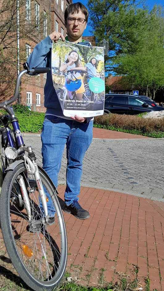 Eine Person steht im Freien auf einem gepflasterten Platz und hält ein großes, leicht geknicktes Plakat zum Stadtradeln in die Kamera. Sie trägt eine blaue Jeans, dunkle Schuhe sowie einen blau-grünen Hoodie unter einer Jacke und hat eine Brille. Neben ihr steht ein Fahrrad, dessen Vorderrad und Lenker im Vordergrund sichtbar sind. Das Plakat zeigt zwei lachende Kinder mit Fahrrädern in warmem Sonnenlicht. Dominierende Farben sind Grün- und Gelbtöne im Hintergrund sowie Blau beim Fahrradhelm. Ein grüner Kreis hebt einen Hinweis zur App hervor. Im Hintergrund des Fotos sind ein rotes Backsteingebäude, ein parkendes dunkles Auto, Bäume mit frischem Grün und ein blauer Himmel zu sehen. Das Licht ist hell und sonnig, die Stimmung insgesamt freundlich, aktiv und einladend.