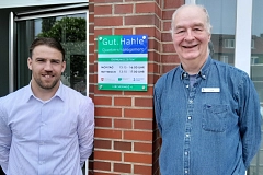 Zwei Männer stehen lächelnd vor dem Eingang des Quartiersmanagements „Gut.Hahle“. Zwischen ihnen befindet sich an einer roten Backsteinwand ein blau-grün-weißes Schild mit den Öffnungszeiten der Einrichtung. Der jüngere Mann links trägt ein hell gestreiftes Hemd, der ältere Mann rechts ein blaues Jeanshemd mit Namensschild. Im Hintergrund sind Fensterfronten, ein geöffneter Eingang sowie Wohnhäuser und Bäume zu sehen. Die Farben wirken freundlich und natürlich, die Stimmung offen und einladend.
