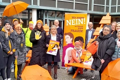 Orange Days 2025 Vor dem Rathaus der Hansestadt Stade steht eine Gruppe von rund 20 Personen unterschiedlichen Alters und Geschlechts zusammen. Sie beteiligen sich an der Aktion „Orange Days“ gegen Gewalt an Frauen. Viele tragen orangefarbene Kleidungsstücke wie Mützen, Schals oder Jacken – Orange ist die Aktionsfarbe. Einige halten orangefarbene Schirme oder Plakate mit dem Slogan „Wir sagen NEIN! zu Gewalt gegen Frauen“. Im Zentrum steht eine orangefarbene Sitzbank mit einer Person, die knieend ein Plakat präsentiert. Hinter ihr ein großes, auffälliges Banner mit illustrierten Frauenporträts. Die Stimmung ist solidarisch und engagiert. Der Hintergrund zeigt ein modernes Gebäude mit großen Fenstern und Backsteinfassade. Ein kleiner grauer Hund sitzt im Vordergrund.