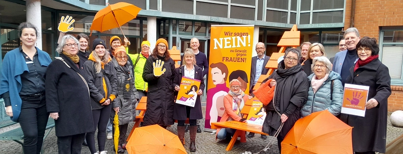 Vor dem Rathaus der Hansestadt Stade steht eine Gruppe von rund 20 Personen unterschiedlichen Alters und Geschlechts zusammen. Sie beteiligen sich an der Aktion „Orange Days“ gegen Gewalt an Frauen. Viele tragen orangefarbene Kleidungsstücke wie Mützen, Schals oder Jacken – Orange ist die Aktionsfarbe. Einige halten orangefarbene Schirme oder Plakate mit dem Slogan „Wir sagen NEIN! zu Gewalt gegen Frauen“. Im Zentrum steht eine orangefarbene Sitzbank mit einer Person, die knieend ein Plakat präsentiert. Hinter ihr ein großes, auffälliges Banner mit illustrierten Frauenporträts. Die Stimmung ist solidarisch und engagiert. Der Hintergrund zeigt ein modernes Gebäude mit großen Fenstern und Backsteinfassade. Ein kleiner grauer Hund sitzt im Vordergrund.