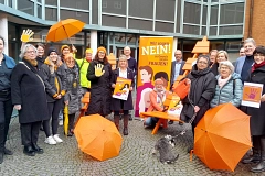Vor dem Rathaus der Hansestadt Stade steht eine Gruppe von rund 20 Personen unterschiedlichen Alters und Geschlechts zusammen. Sie beteiligen sich an der Aktion „Orange Days“ gegen Gewalt an Frauen. Viele tragen orangefarbene Kleidungsstücke wie Mützen, Schals oder Jacken – Orange ist die Aktionsfarbe. Einige halten orangefarbene Schirme oder Plakate mit dem Slogan „Wir sagen NEIN! zu Gewalt gegen Frauen“. Im Zentrum steht eine orangefarbene Sitzbank mit einer Person, die knieend ein Plakat präsentiert. Hinter ihr ein großes, auffälliges Banner mit illustrierten Frauenporträts. Die Stimmung ist solidarisch und engagiert. Der Hintergrund zeigt ein modernes Gebäude mit großen Fenstern und Backsteinfassade. Ein kleiner grauer Hund sitzt im Vordergrund.