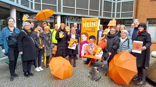 Vor dem Rathaus der Hansestadt Stade steht eine Gruppe von rund 20 Personen unterschiedlichen Alters und Geschlechts zusammen. Sie beteiligen sich an der Aktion „Orange Days“ gegen Gewalt an Frauen. Viele tragen orangefarbene Kleidungsstücke wie Mützen, Schals oder Jacken – Orange ist die Aktionsfarbe. Einige halten orangefarbene Schirme oder Plakate mit dem Slogan „Wir sagen NEIN! zu Gewalt gegen Frauen“. Im Zentrum steht eine orangefarbene Sitzbank mit einer Person, die knieend ein Plakat präsentiert. Hinter ihr ein großes, auffälliges Banner mit illustrierten Frauenporträts. Die Stimmung ist solidarisch und engagiert. Der Hintergrund zeigt ein modernes Gebäude mit großen Fenstern und Backsteinfassade. Ein kleiner grauer Hund sitzt im Vordergrund.