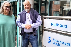 Zwei lächelnde Personen stehen vor dem Eingang des Rathauses der Hansestadt Stade. Rechts im Bild ist ein großes Schild mit der Aufschrift „Rathaus – Hansestadt Stade“ zu sehen. Die Frau trägt eine mintgrüne Tunika und Brille, der Mann ein helles Hemd mit Pullover über den Schultern und hält die Hände zusammen.
