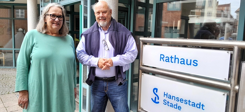 Zwei lächelnde Personen stehen vor dem Eingang des Rathauses der Hansestadt Stade. Rechts im Bild ist ein großes Schild mit der Aufschrift „Rathaus – Hansestadt Stade“ zu sehen. Die Frau trägt eine mintgrüne Tunika und Brille, der Mann ein helles Hemd mit Pullover über den Schultern und hält die Hände zusammen.