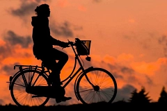 Eine Person fährt bei Sonnenuntergang auf einem Fahrrad. Die Szene ist als dunkle Silhouette vor einem orange gefärbten Himmel mit Wolken zu sehen.