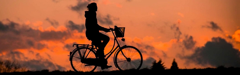 Eine Person fährt bei Sonnenuntergang auf einem Fahrrad. Die Szene ist als dunkle Silhouette vor einem orange gefärbten Himmel mit Wolken zu sehen.