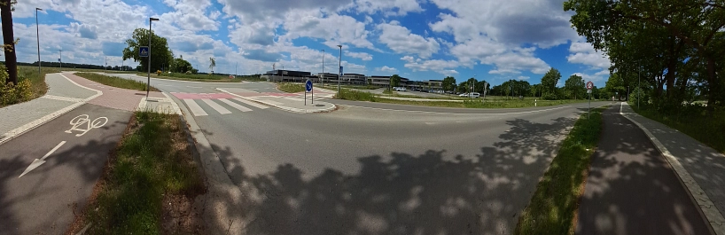 Panoramaaufnahme eines modernen Kreisverkehrs mit neu angelegtem Radweg und Zebrastreifen an der Harsefelder Straße in Stade. Links und rechts verlaufen separate Geh- und Radwege, im Hintergrund moderne Gebäude, Autos und offene Landschaft.