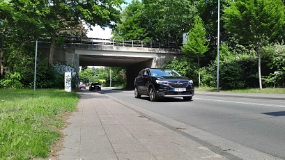 PI - B73-Brückenneubau geplant Straßenszene mit mehreren Autos, die unter einer Brücke hindurchfahren. Die Brücke ist aus Beton und mit Graffiti besprüht. Links und rechts säumen grüne Bäume und Büsche die Straße. Ein schwarzer SUV fährt auf der rechten Spur, im Hintergrund sind weitere Fahrzeuge und ein gelbes Verkehrsschild zu sehen.