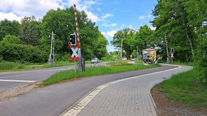 Ein Bahnübergang an der Harsefelder Straße in Stade. Zu sehen sind eine Fahrbahn, ein separater Rad- und Gehweg mit neuem Pflaster sowie Schranken und Signalanlagen. Im Hintergrund ein Auto und dichte, grüne Vegetation.