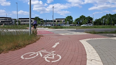 Ein neu gestalteter Fahrradweg mit rotem Pflaster und Richtungspfeil führt zu einem Zebrastreifen an einem Kreisverkehr. Eine Person fährt mit dem Fahrrad, im Hintergrund sind Gebäude und Parkplatz des Bildungscampus Riensförde zu sehen.