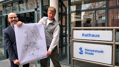 Zwei Männer stehen vor dem Eingang des Rathauses der Hansestadt Stade. Sie halten gemeinsam einen großen Stadtplan in den Händen, der die kommunale Wärmeplanung zeigt. Rechts im Bild ist ein Schild mit der Aufschrift „Rathaus – Hansestadt Stade“ zu sehen.