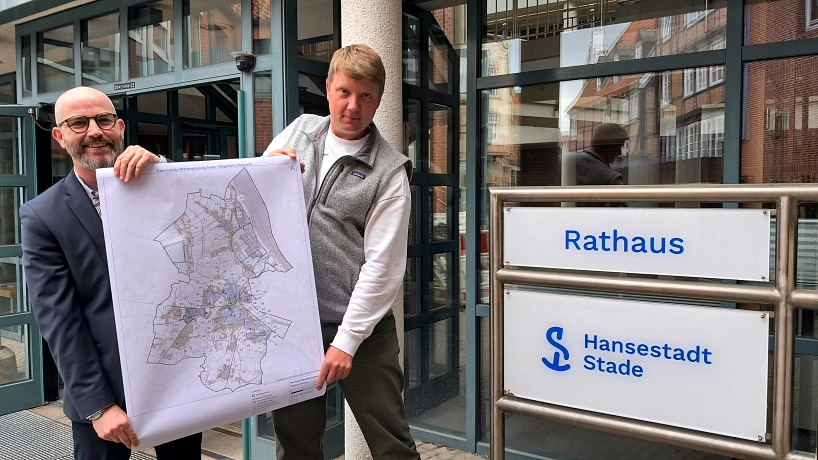 PI - Kommunaler Wärmeplan veröffentlicht Zwei Männer stehen vor dem Eingang des Rathauses der Hansestadt Stade. Sie halten gemeinsam einen großen Stadtplan in den Händen, der die kommunale Wärmeplanung zeigt. Rechts im Bild ist ein Schild mit der Aufschrift „Rathaus – Hansestadt Stade“ zu sehen. © Hansestadt Stade