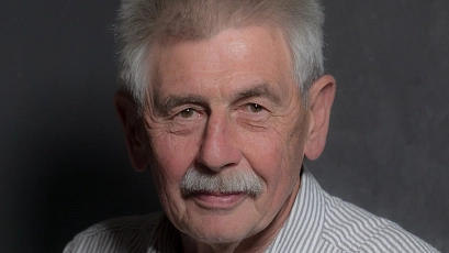 Ein älterer Mann mit grauen Haaren und Schnurrbart blickt freundlich in die Kamera. Er trägt ein hell gestreiftes Hemd und sitzt vor einem neutralen, dunklen Hintergrund.