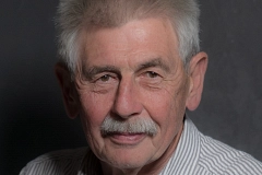 Portrait Ernst Ryll Ein älterer Mann mit grauen Haaren und Schnurrbart blickt freundlich in die Kamera. Er trägt ein hell gestreiftes Hemd und sitzt vor einem neutralen, dunklen Hintergrund.