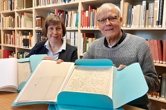 Zwei Personen sitzen an einem Holztisch in einem Archivraum und halten aufgeschlagene historische Dokumente in hellblauen Mappen in die Kamera. Hinter ihnen stehen hohe Bücherregale mit dicht aneinandergereihten Büchern in verschiedenen Farben wie Beige, Rot und Blau. Die Frau trägt ein dunkles Sakko über einer hellen Bluse, der Mann einen grauen Strickpullover und eine Brille. Beide lächeln freundlich und wirken zugewandt. Die Dokumente zeigen vergilbtes Papier mit handschriftlichen Einträgen. Das warme Licht und die ruhige Umgebung vermitteln eine konzentrierte, zugleich einladende Arbeitsatmosphäre.