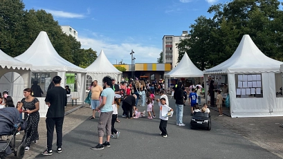 Menschen unterschiedlicher Altersgruppen besuchen ein Sommerfest im Stadtteil. Zwischen weißen Pagodenzelten bewegen sich Erwachsene und Kinder, einige Kinder spielen oder fahren mit einem Spielzeugauto. Im Hintergrund sind Bäume und ein mehrstöckiges Wohnhaus zu sehen. Die Atmosphäre ist lebendig und einladend.
