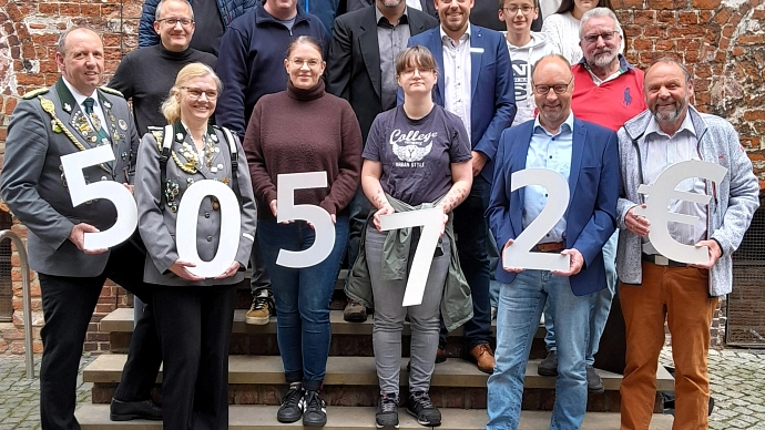 Gruppenfoto vor einem historischen Backsteingebäude mit großem Rundbogenportal und türkisfarbener Holztür. Rund 30 Personen unterschiedlichen Alters stehen auf einer Treppe und schauen in die Kamera. Die Stimmung wirkt freundlich und feierlich. Im Vordergrund halten mehrere Personen große weiße Zeichen mit der Aufschrift „50 572 €“. Die Kleidung reicht von Anzügen und Sakkos über Freizeitkleidung bis hin zu Schützenuniformen in Grau mit silbernen Abzeichen. Der Hintergrund wird von roten Backsteinen, hellen Sandsteinelementen und gepflastertem Boden geprägt.