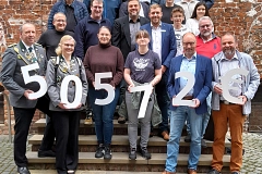 Gruppenfoto vor einem historischen Backsteingebäude mit großem Rundbogenportal und türkisfarbener Holztür. Rund 30 Personen unterschiedlichen Alters stehen auf einer Treppe und schauen in die Kamera. Die Stimmung wirkt freundlich und feierlich. Im Vordergrund halten mehrere Personen große weiße Zeichen mit der Aufschrift „50 572 €“. Die Kleidung reicht von Anzügen und Sakkos über Freizeitkleidung bis hin zu Schützenuniformen in Grau mit silbernen Abzeichen. Der Hintergrund wird von roten Backsteinen, hellen Sandsteinelementen und gepflastertem Boden geprägt.