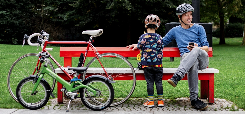 Ein Mann und sein Sohn machen es sich bei einer Parkbank bequem. Neben ihnen stehen, an die Bank gelehnt, ihre Fahrräder