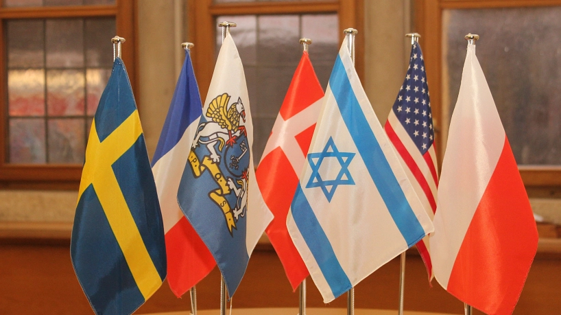 Sieben kleine Tischflaggen stehen nebeneinander aufgereiht in einem Innenraum mit großen Fenstern im Hintergrund. Von links nach rechts sind folgende Fahnen zu sehen: Schweden, Frankreich, das Stader Wappen, Dänemark, Israel, USA und Polen. Die Szene wirkt feierlich und symbolisiert internationale Partnerschaften.